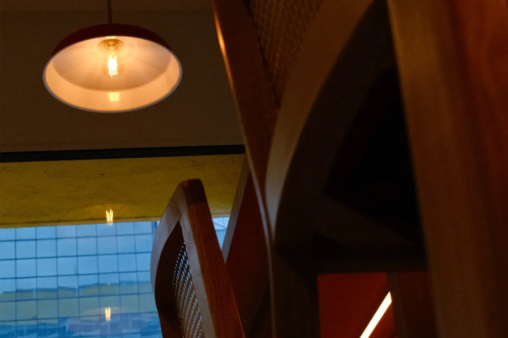A glowing ceiling lamp illuminates a dim, angular interior with blue grid windows and a narrow bright doorway, creating a quiet, moody atmosphere.