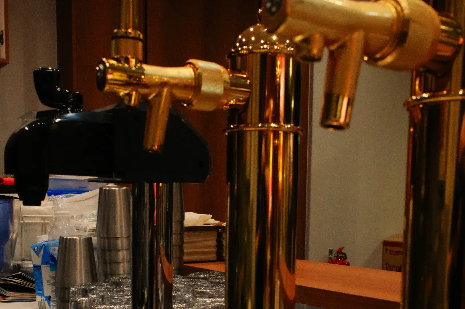 Polished brass beer taps dominate a warmly lit bar, with stacked glasses and a black dispenser behind them, creating a cozy pub atmosphere.