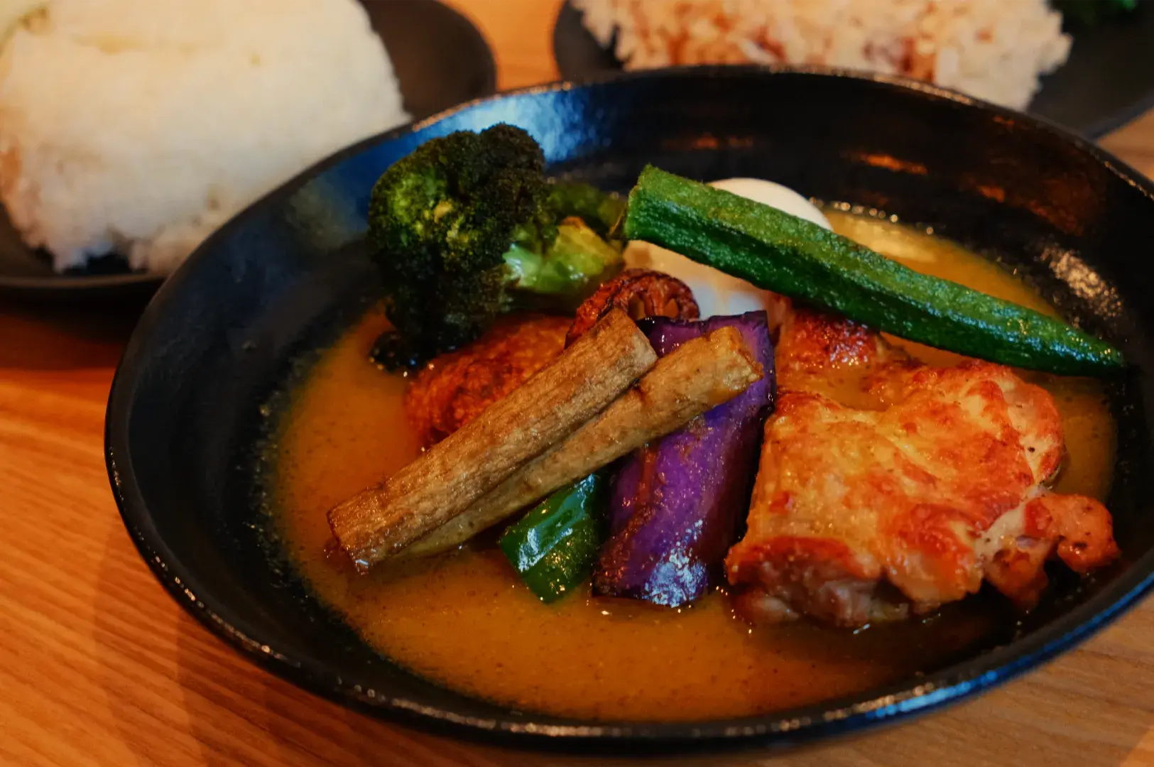 A black bowl of golden curry holds browned chicken and colorful vegetables, including zucchini, eggplant and broccoli, with rice served alongside.