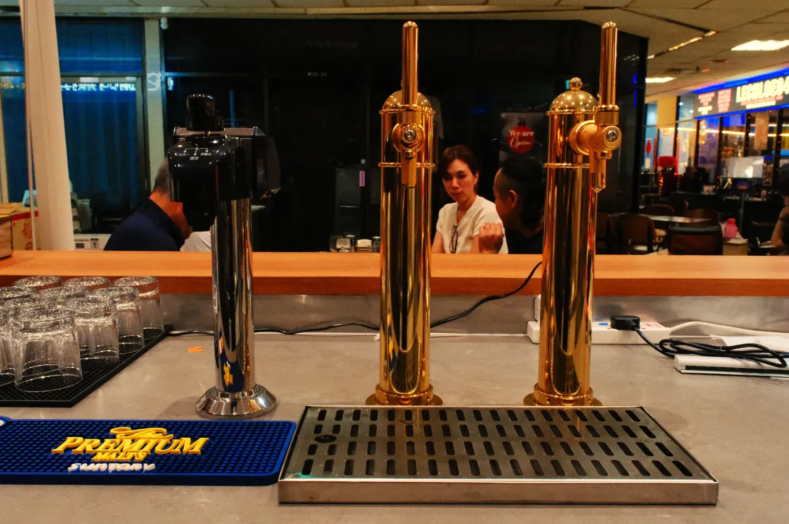 Three polished brass beer taps stand on a bar before a mirror, with glasses and a blue bar mat nearby; staff converse in the background, creating a busy pub atmosphere.