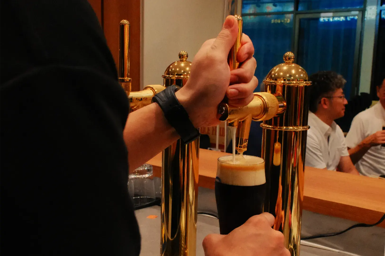 A bartender pulls a dark stout with a creamy tan head from a polished brass tap, while a colleague works behind the bar. Warm, busy pub atmosphere.