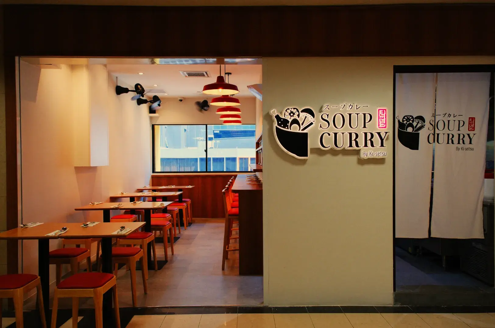 Warmly lit soup curry restaurant with wooden tables, red chairs, a large window, and matching bowl logos on the wall and doorway curtain.