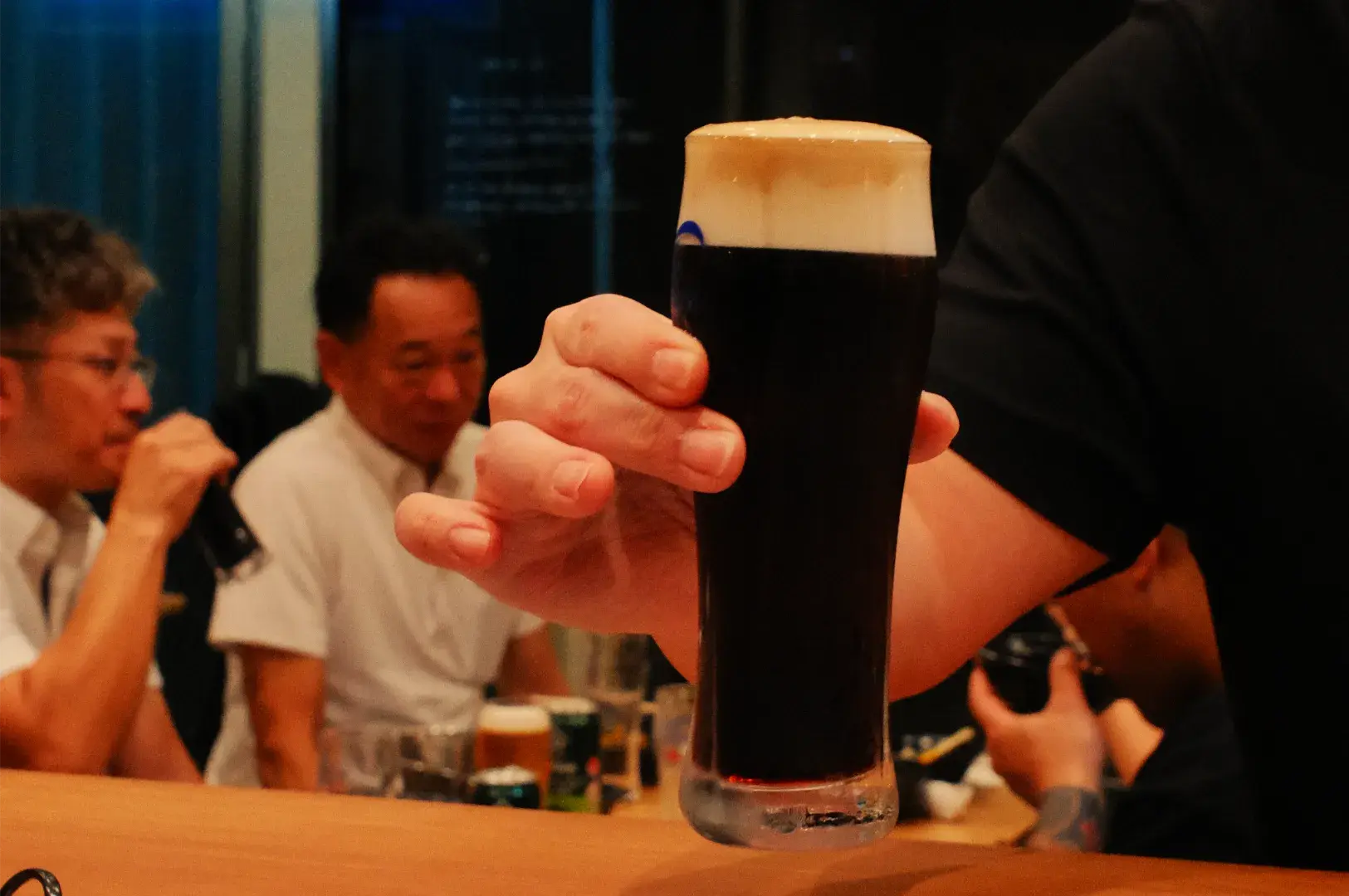 A hand presents a pint of dark stout topped with creamy tan foam in a warmly lit bar, while diners chat in the background; a convivial scene.
