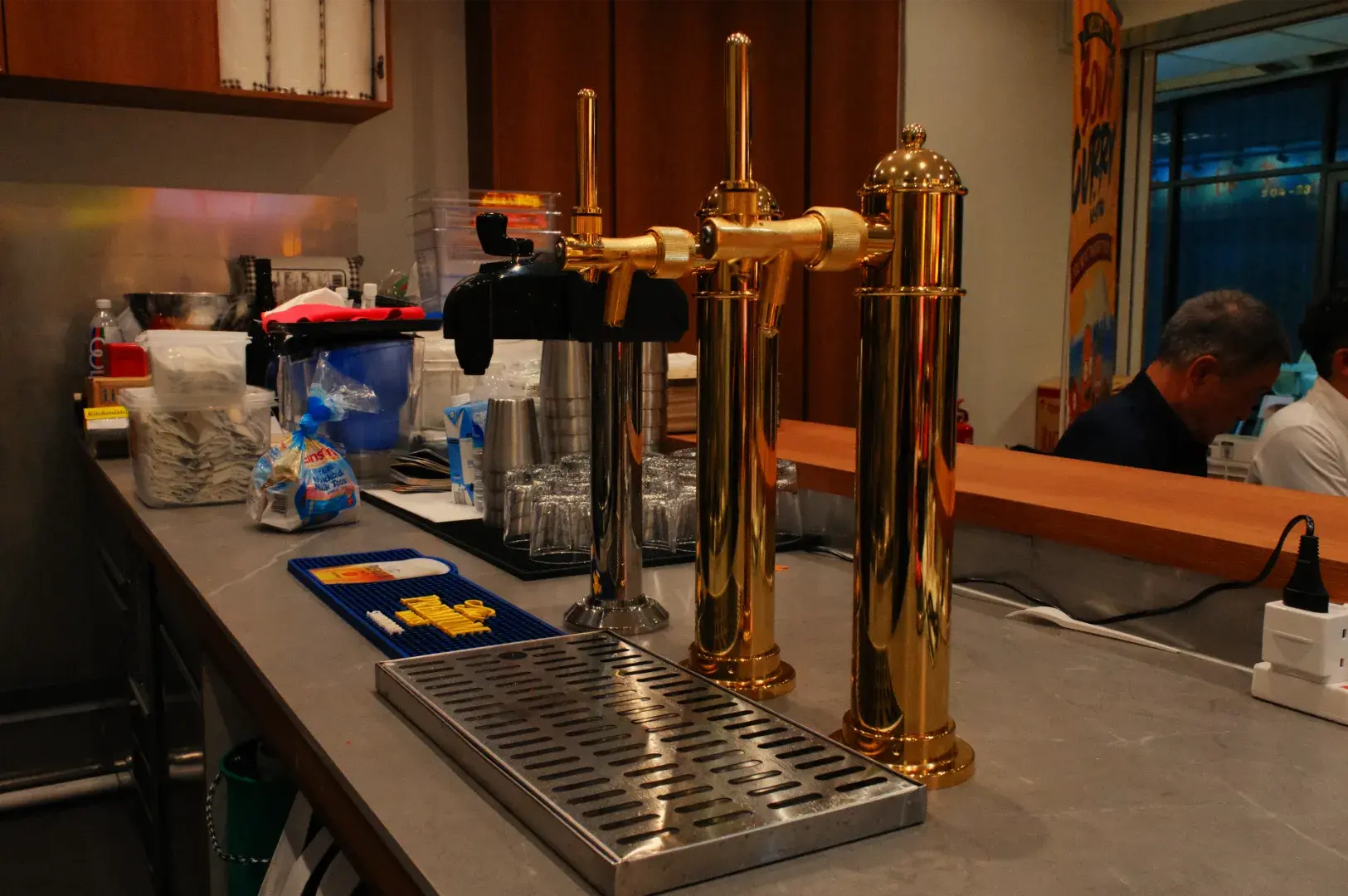Three polished brass beer taps stand on a caf&eacute; counter beside a metal drip tray, with a barista and customer in the background, creating a warm, busy pub atmosphere.