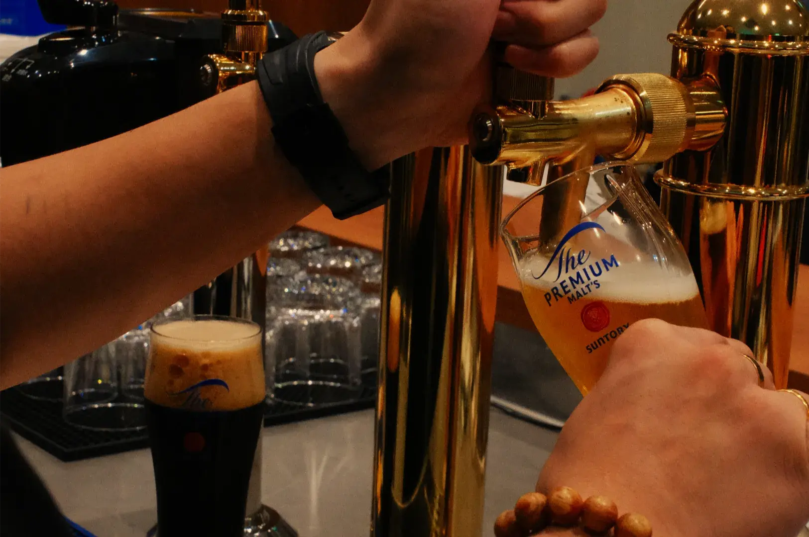 A bartender pulls a brass beer tap, pouring golden beer into a tilted branded glass beside a dark stout, creating a warm, lively bar scene.