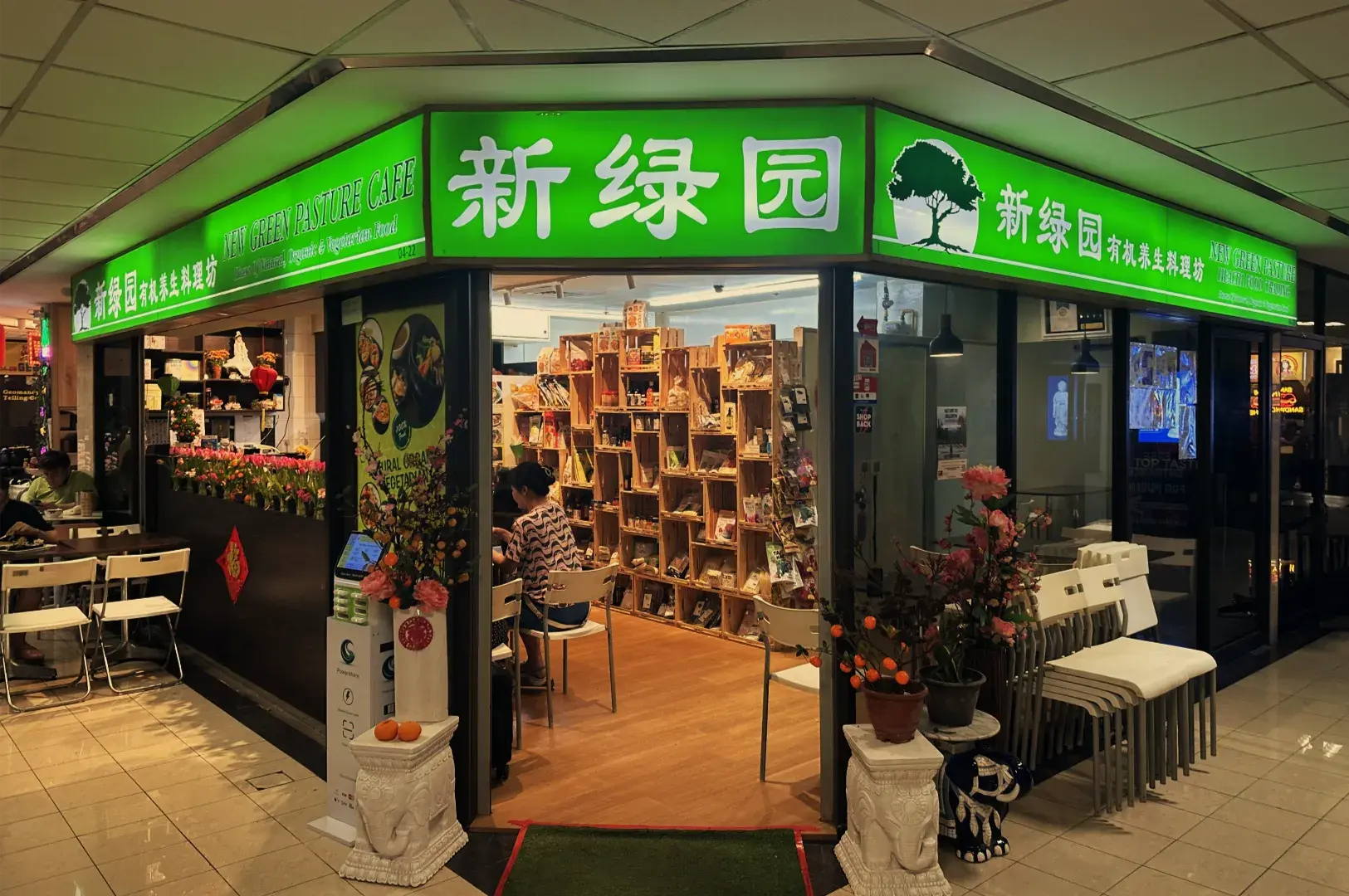 A warmly lit Chinese tea shop with bright green signage, wooden tea shelves, potted flowers, and outdoor seating in a busy indoor market.