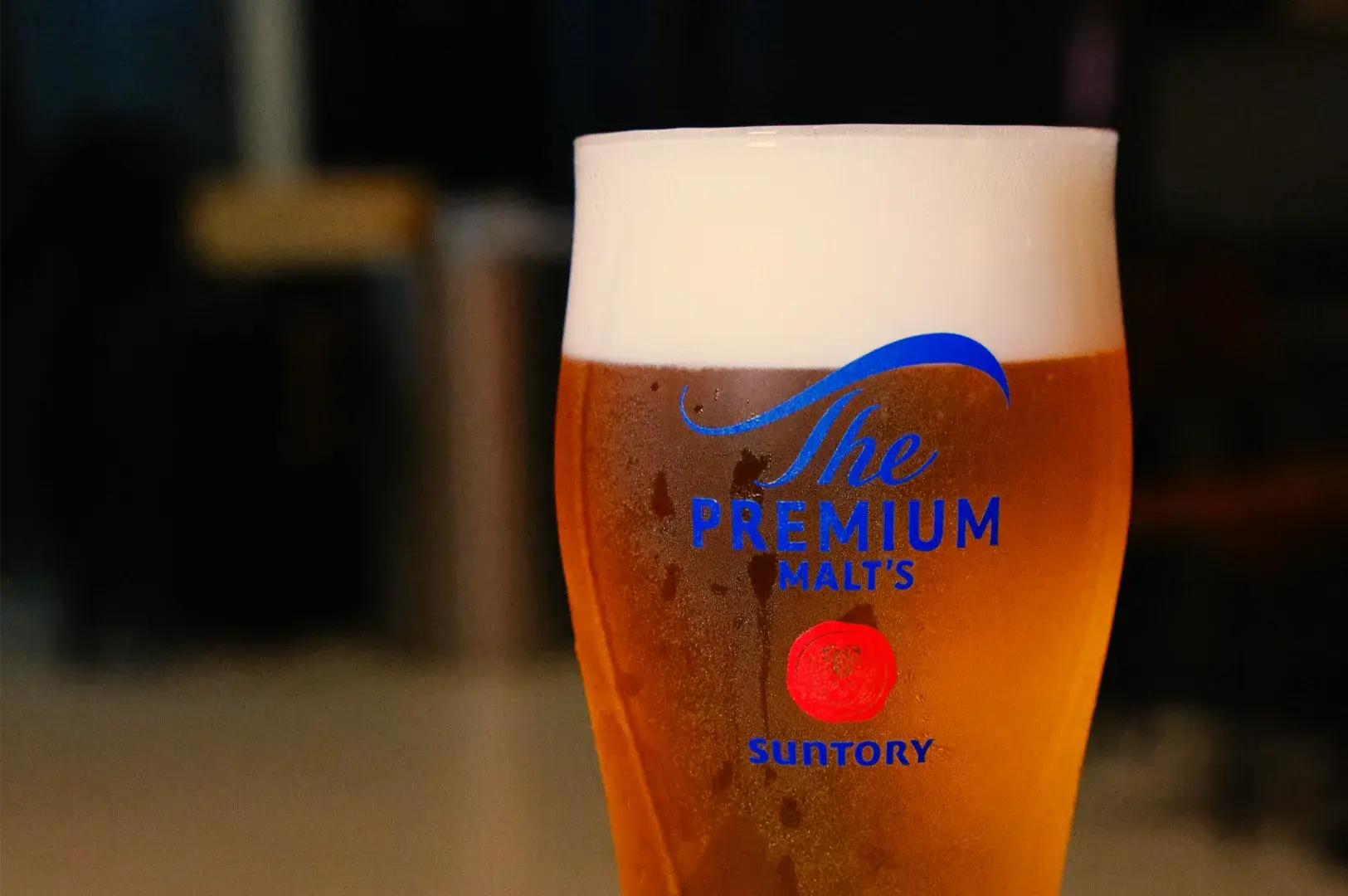 A chilled glass of amber Suntory Premium Malt&rsquo;s beer with a thick white foam head sits against a softly blurred bar background, inviting and warm.
