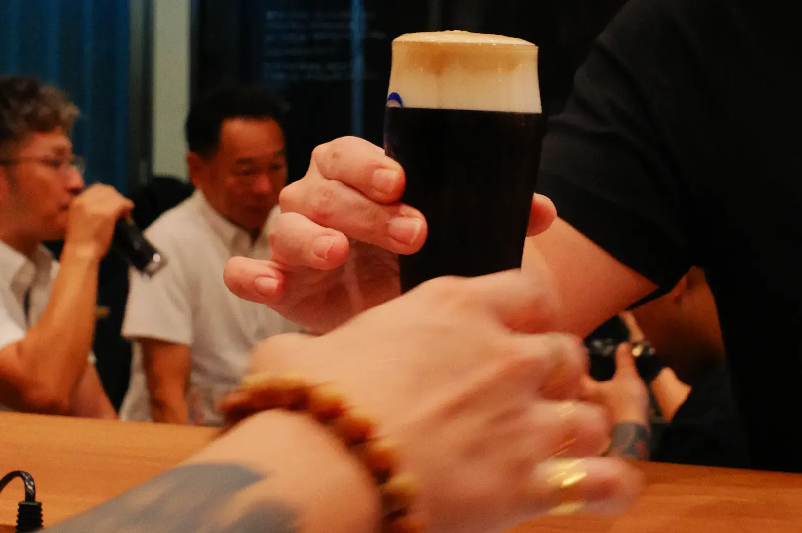 A hand raises a dark stout with a creamy foam head at a lively bar, while patrons talk and drink in the warm, convivial atmosphere.