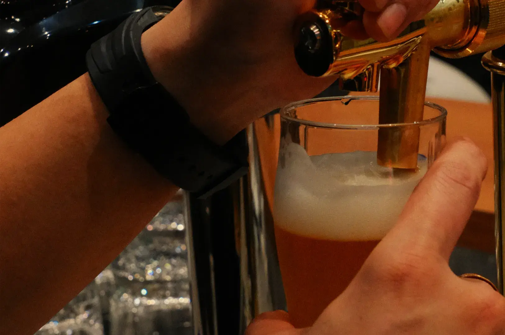 Close-up of hands pouring amber beer from a tap into a glass, forming a foamy head in a dimly lit bar; a relaxed, social mood.