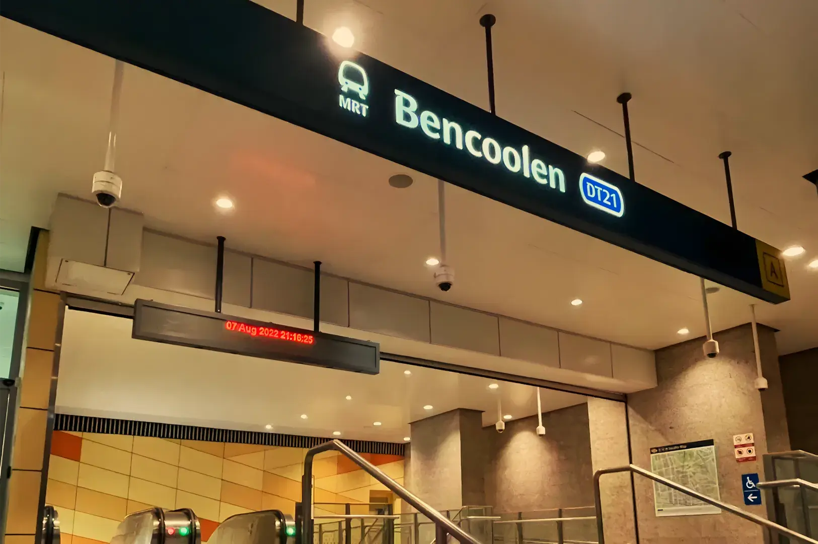 Warmly lit interior of Singapore&rsquo;s Bencoolen MRT station, with the illuminated station name and blue train-line marker above ticket gates and stairs.