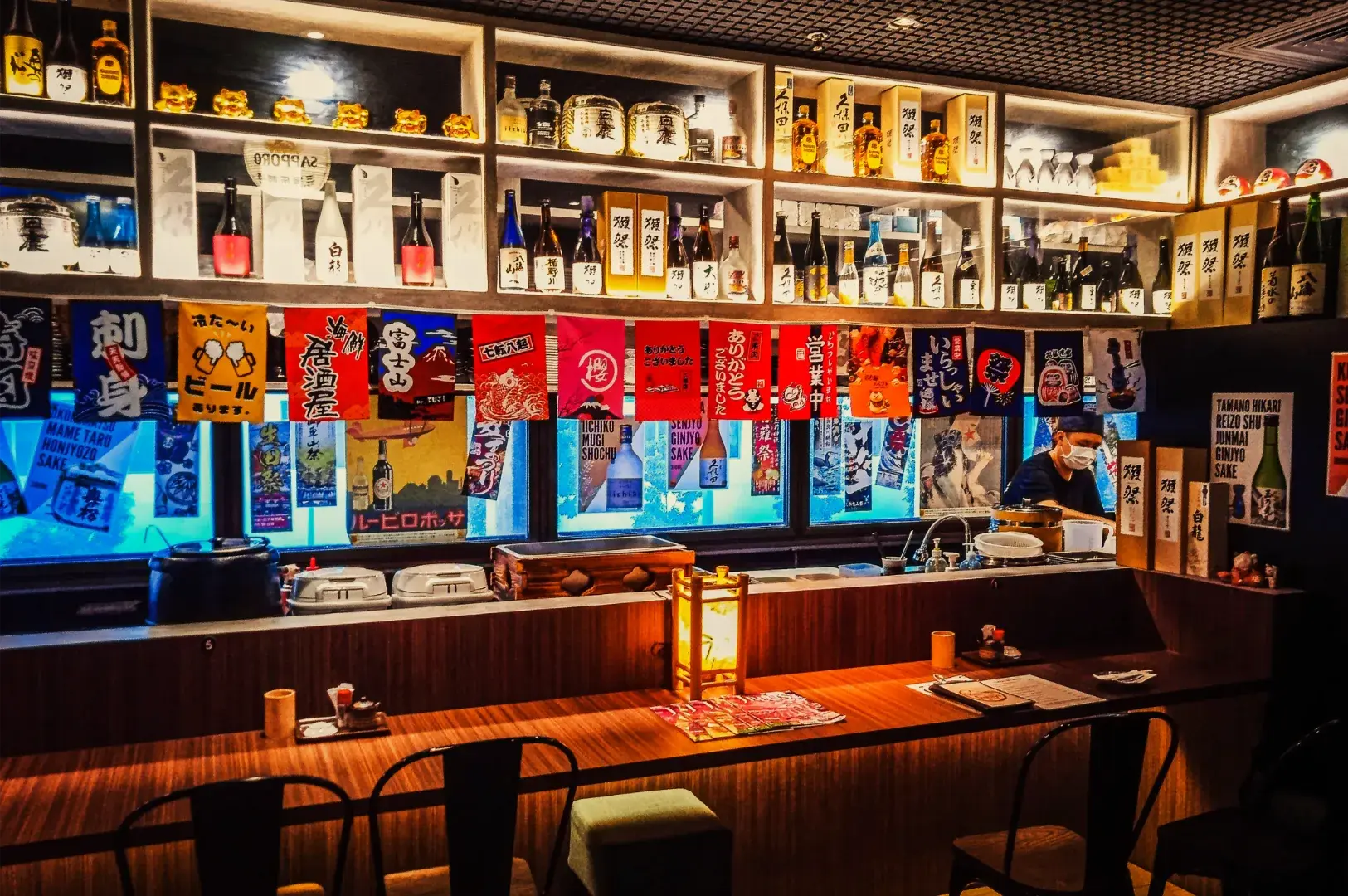 A cozy Japanese izakaya counter lined with sake bottles and colorful menu banners, with a glowing lantern and a masked cook behind the bar.