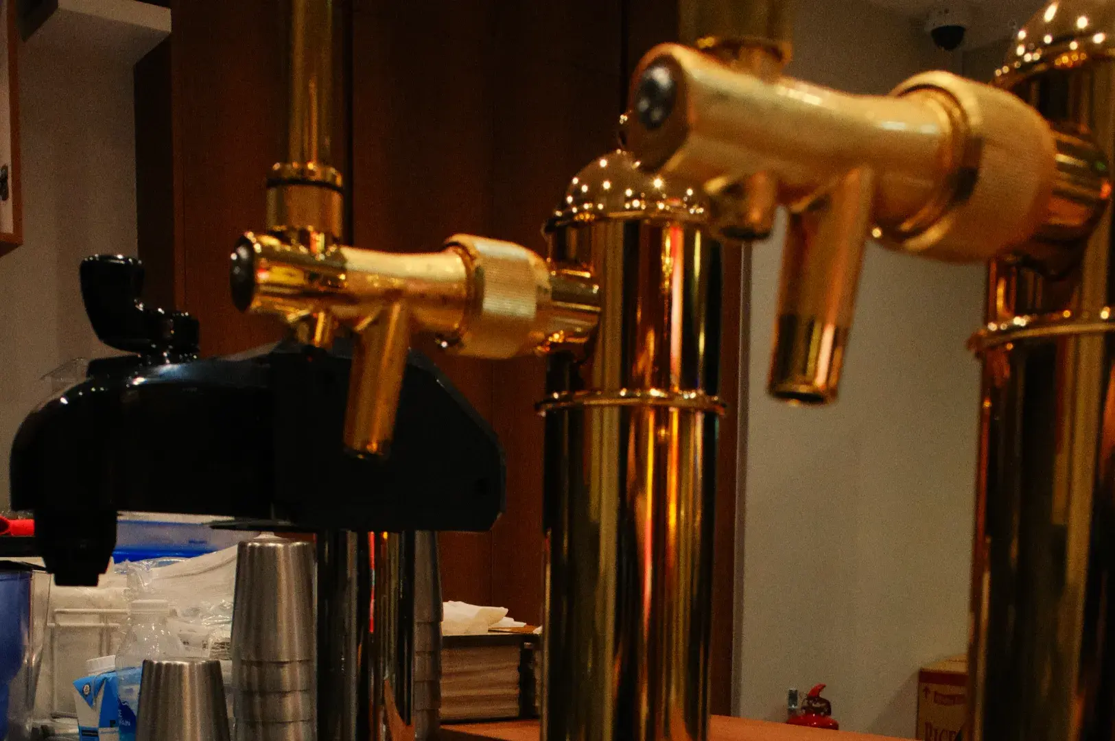 Polished brass beer taps and a black dispenser stand at a bar, their reflective metal glowing in the warm, dim atmosphere of a restaurant.