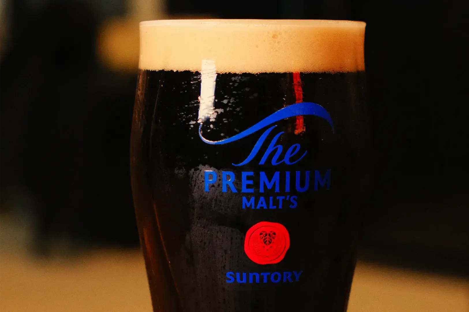 A glass of dark Suntory The Premium Malt&rsquo;s beer with a thick golden foam head and blue logo, warmly lit against a blurred background.