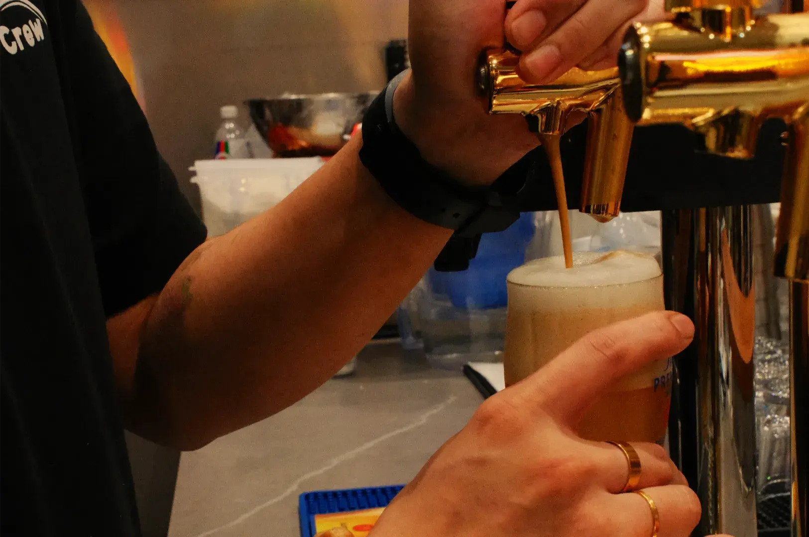 Close-up of a bartender&rsquo;s hands pulling a polished brass tap to pour golden beer into a pint glass, building a creamy foam head in a warm bar.