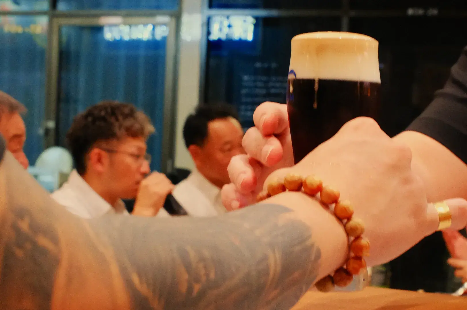 A tattooed arm and several hands raise a dark beer with a creamy head in a crowded bar, as blurred patrons share a warm, celebratory toast.