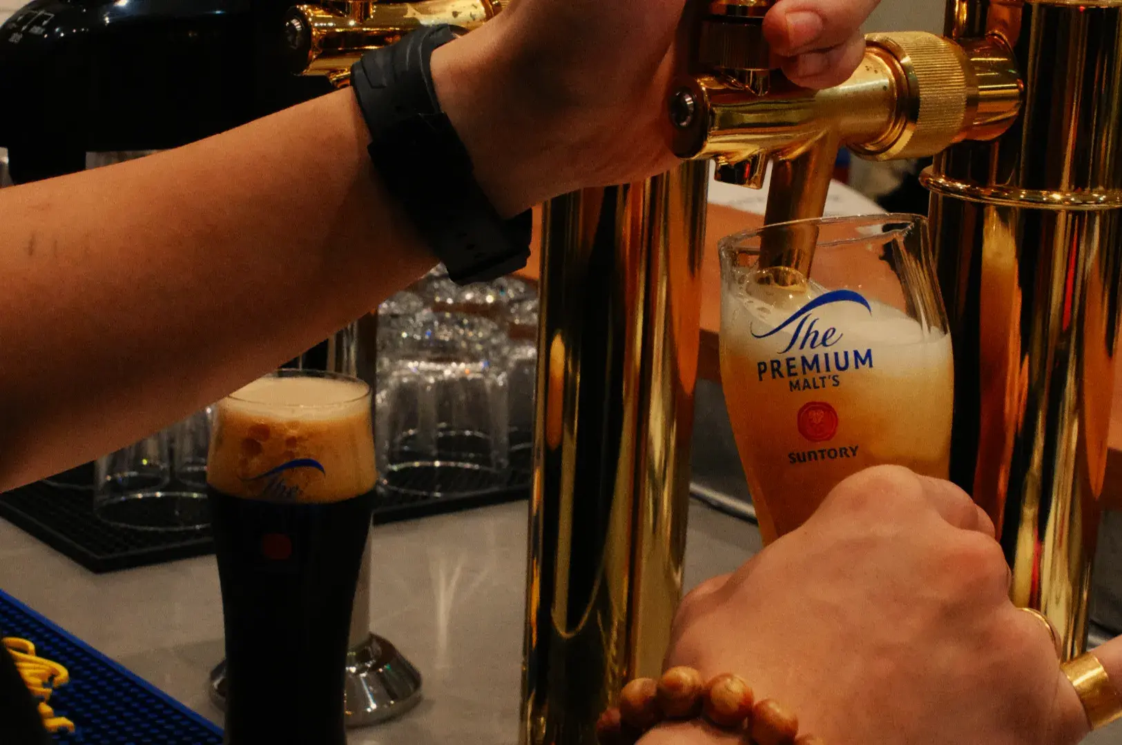 A bartender pulls a polished brass tap to pour Suntory Premium Malt&rsquo;s beer into a tilted glass, amid glowing bar taps and a warm, busy atmosphere.